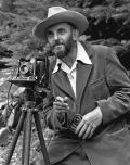 Der Fotograf Ansel Adams mit Hut neben einer Kamera auf einem Stativ
