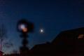 Mondfotografie – den Mond über den Hausdächern fotografieren Teilbeleuchteter Mond über Hausdächern mit unscharf abgebildeter Kamera auf einem Stativ im Vordergrund.